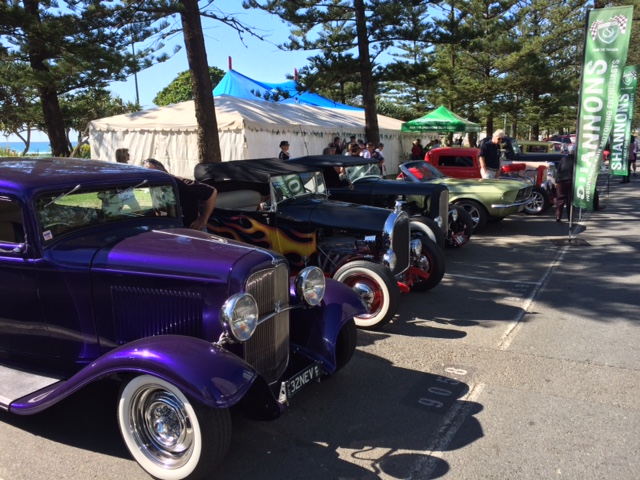 A row of colorful vintage cars is parked at an outdoor car show. Tents and trees are in the background under a clear blue sky, creating a lively atmosphere.